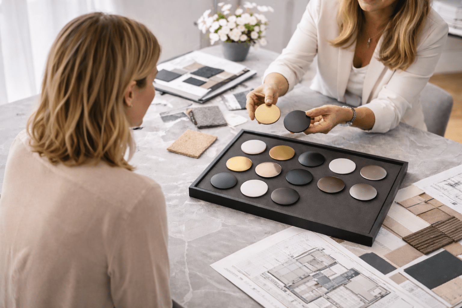 Overhead / top-down shot of a design table. A single female client and an interior designer are seated opposite each other (only arms and torsos visible). On the table is a tray of small circular metal and matte finish samples in brass, bronze, black and white, alongside fabric swatches, stone samples and architectural drawings.